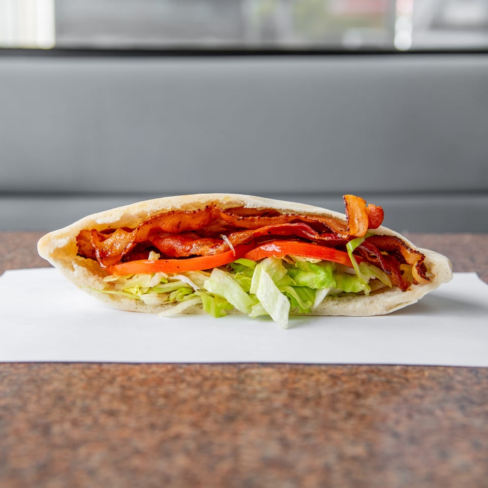 Bacon-filled pita sandwich with lettuce and tomato, served on white paper atop a wooden table.