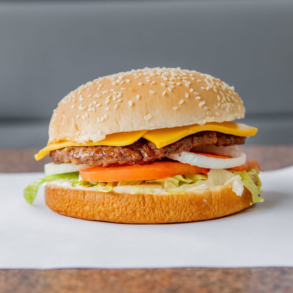 Cheeseburger with sesame bun, beef patty, lettuce, tomato, onion, sauce, and melted cheese on a white plate.